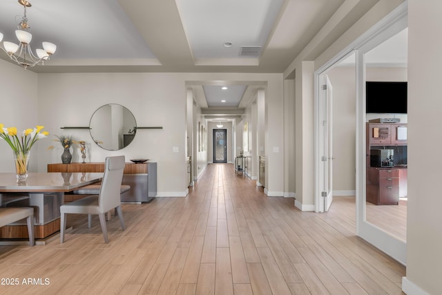 hall with light wood finished floors, visible vents, baseboards, a tray ceiling, and an inviting chandelier