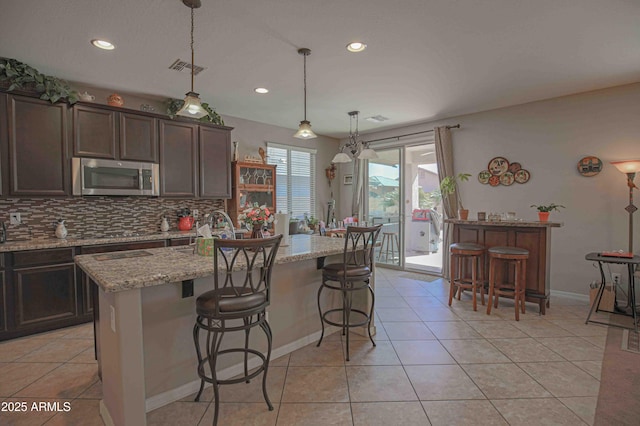 kitchen featuring tasteful backsplash, a kitchen island with sink, stainless steel microwave, and light tile patterned floors