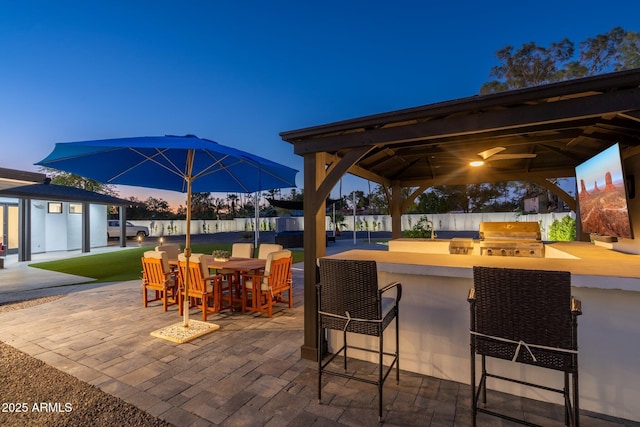 patio terrace at dusk featuring fence, a grill, an outdoor kitchen, a gazebo, and a bar
