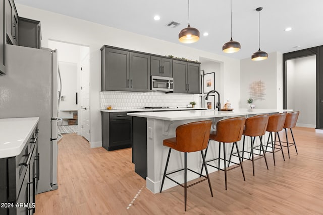 kitchen with light wood-type flooring, a kitchen island with sink, hanging light fixtures, and decorative backsplash