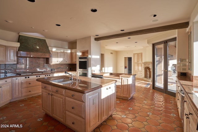 kitchen with sink, an island with sink, decorative backsplash, custom exhaust hood, and appliances with stainless steel finishes