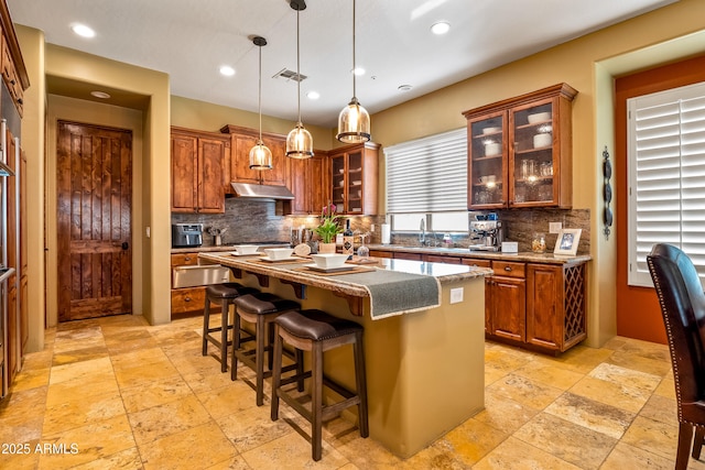 kitchen with under cabinet range hood, stone tile floors, a kitchen island, hanging light fixtures, and glass insert cabinets