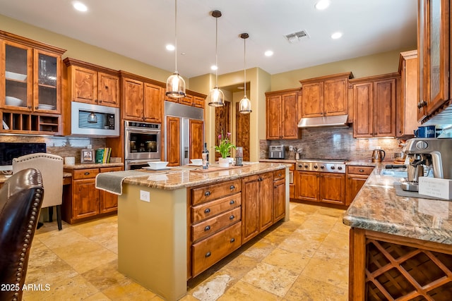 kitchen featuring built in appliances, light stone counters, under cabinet range hood, hanging light fixtures, and an island with sink