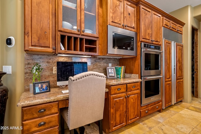 kitchen featuring glass insert cabinets, light stone counters, built in appliances, built in desk, and backsplash