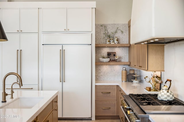 kitchen with a sink, paneled built in fridge, light countertops, wall chimney exhaust hood, and stainless steel range with gas stovetop