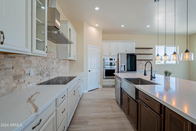 kitchen with appliances with stainless steel finishes, white cabinetry, wall chimney exhaust hood, pendant lighting, and light hardwood / wood-style flooring