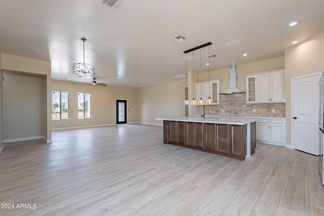 kitchen with wall chimney exhaust hood, an island with sink, hanging light fixtures, white cabinetry, and light hardwood / wood-style floors