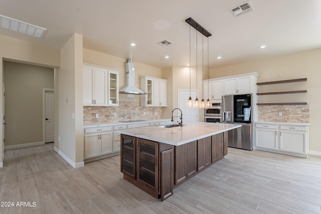 kitchen with wall chimney range hood, hanging light fixtures, a kitchen island with sink, sink, and stainless steel appliances