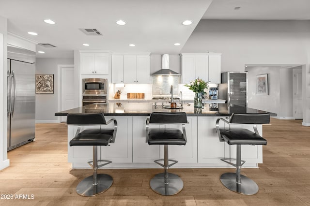 kitchen featuring built in appliances, visible vents, white cabinetry, wall chimney exhaust hood, and light wood finished floors