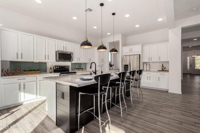 kitchen featuring white cabinetry, wood-type flooring, decorative light fixtures, a large island with sink, and appliances with stainless steel finishes