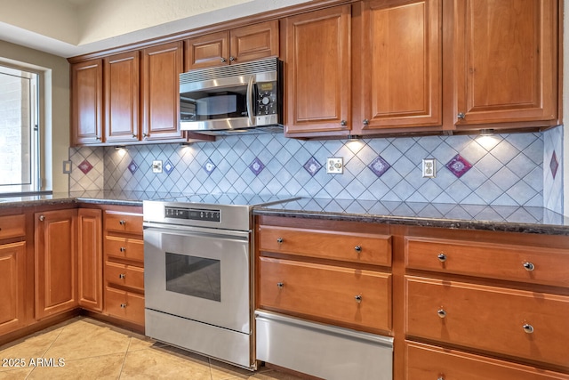 kitchen with light tile patterned floors, stainless steel appliances, decorative backsplash, brown cabinetry, and dark stone countertops