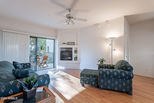 living area featuring a ceiling fan, a glass covered fireplace, baseboards, and wood finished floors