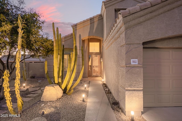 exterior entry at dusk featuring an attached garage, a tile roof, and stucco siding