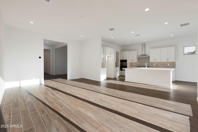 interior space with white cabinets, backsplash, hardwood / wood-style flooring, wall chimney range hood, and a center island with sink