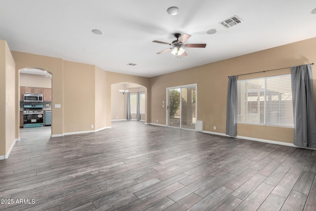 unfurnished living room with arched walkways, ceiling fan with notable chandelier, dark wood-style flooring, visible vents, and baseboards