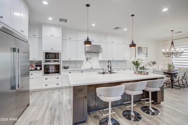 kitchen featuring built in appliances, decorative light fixtures, white cabinetry, and a kitchen island with sink