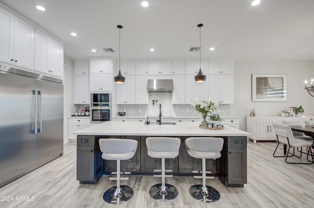 kitchen with wall chimney exhaust hood, stainless steel appliances, a kitchen island with sink, pendant lighting, and white cabinetry