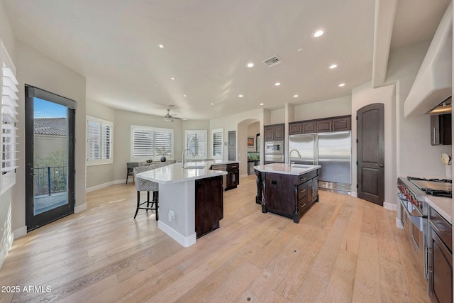 kitchen featuring a center island with sink, visible vents, light countertops, dark brown cabinets, and wall chimney range hood