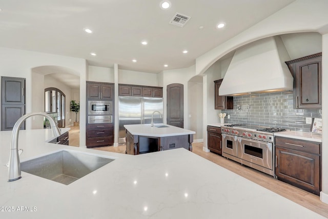 kitchen featuring custom range hood, visible vents, a sink, an island with sink, and built in appliances