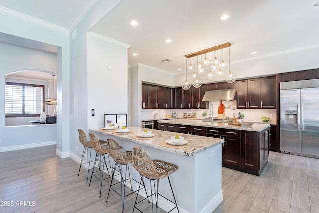 kitchen featuring appliances with stainless steel finishes, ornamental molding, a kitchen island with sink, light wood-type flooring, and under cabinet range hood