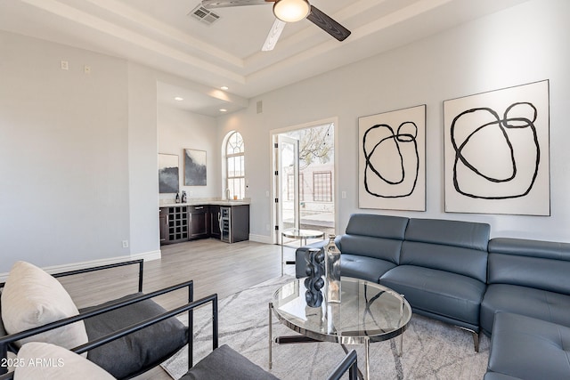 living area with ceiling fan, light wood-style flooring, visible vents, baseboards, and a tray ceiling