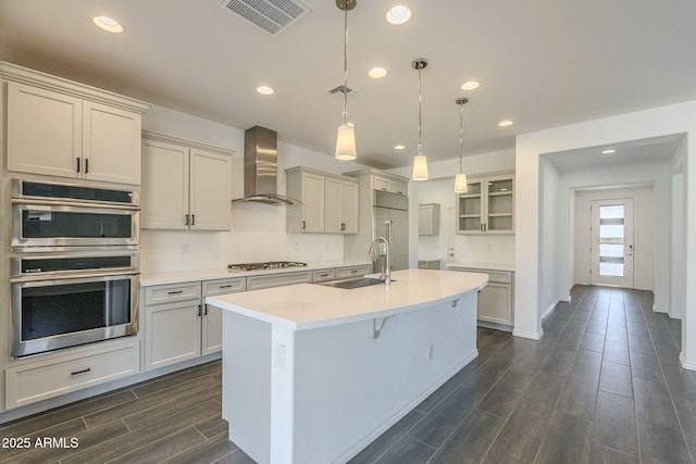 kitchen with decorative light fixtures, wall chimney range hood, stainless steel appliances, sink, and a center island with sink