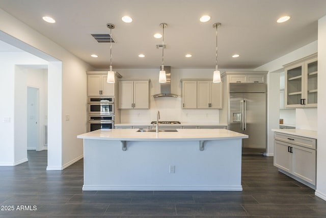 kitchen featuring wall chimney range hood, stainless steel appliances, a kitchen island with sink, and pendant lighting