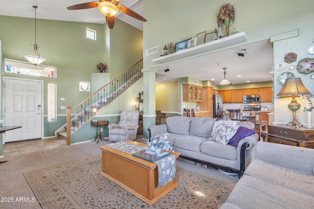 living area featuring light colored carpet, visible vents, stairway, a towering ceiling, and ceiling fan