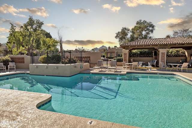 pool at dusk featuring a patio, fence, and a community pool