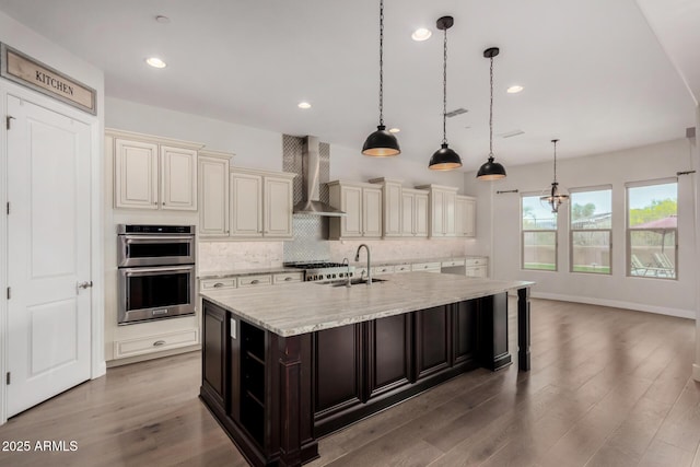 kitchen featuring a sink, backsplash, stainless steel double oven, cream cabinetry, and wall chimney range hood