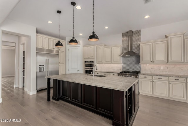kitchen with a sink, a large island with sink, cream cabinets, stainless steel appliances, and wall chimney exhaust hood