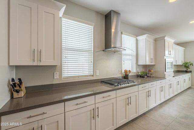 kitchen featuring dark countertops, appliances with stainless steel finishes, light tile patterned flooring, white cabinets, and wall chimney exhaust hood