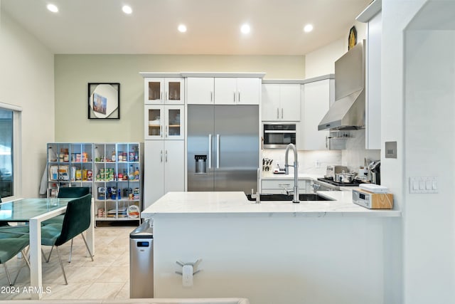 kitchen with kitchen peninsula, wall chimney exhaust hood, stainless steel appliances, sink, and white cabinets