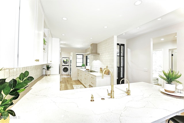 kitchen featuring black electric stovetop, wall chimney range hood, light hardwood / wood-style flooring, decorative backsplash, and stacked washer / drying machine