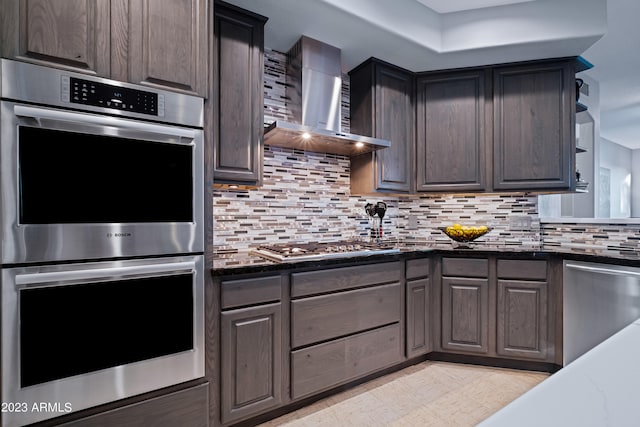 kitchen featuring wall chimney exhaust hood, dark brown cabinets, dark stone countertops, stainless steel appliances, and backsplash