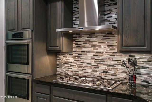 kitchen with wall chimney range hood, backsplash, dark brown cabinets, stainless steel appliances, and dark stone counters