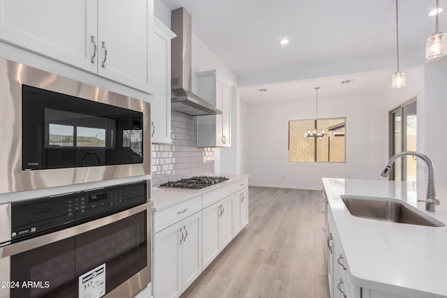 kitchen featuring wall chimney range hood, hanging light fixtures, sink, white cabinets, and appliances with stainless steel finishes