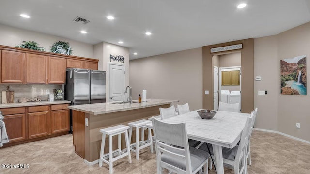 kitchen with washing machine and dryer, a sink, freestanding refrigerator, brown cabinetry, and a center island with sink