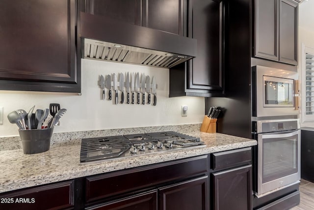 kitchen with dark brown cabinets, wood finished floors, stainless steel appliances, exhaust hood, and light stone counters