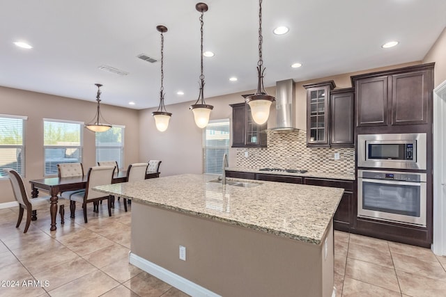 kitchen featuring wall chimney exhaust hood, stainless steel appliances, light stone counters, decorative light fixtures, and a center island with sink