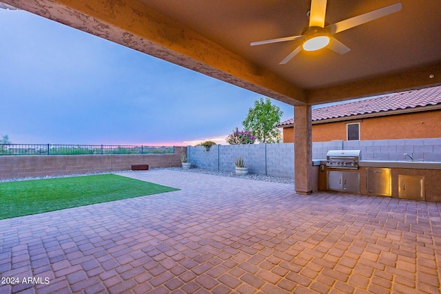 patio terrace at dusk with sink, grilling area, a yard, ceiling fan, and exterior kitchen