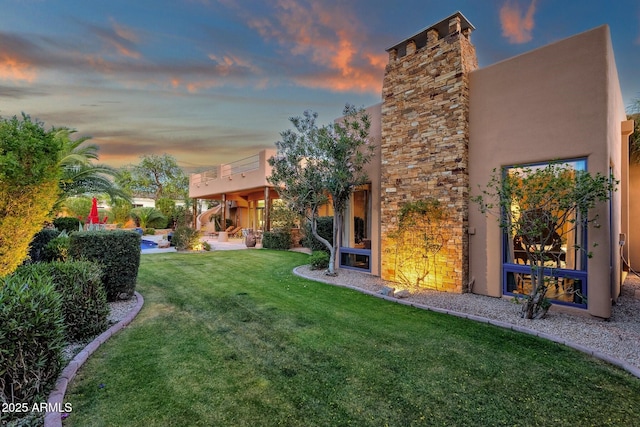 back of property at dusk with stucco siding, a chimney, and a yard
