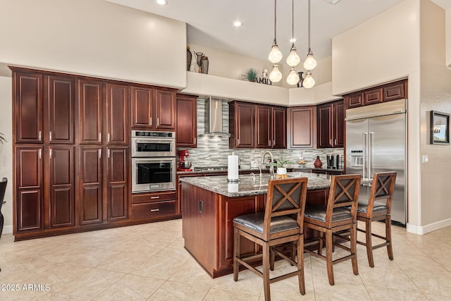 kitchen with appliances with stainless steel finishes, a center island with sink, a kitchen bar, dark stone counters, and wall chimney exhaust hood