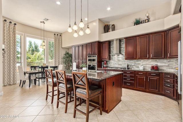 kitchen featuring a breakfast bar, decorative light fixtures, an island with sink, dark stone counters, and wall chimney exhaust hood