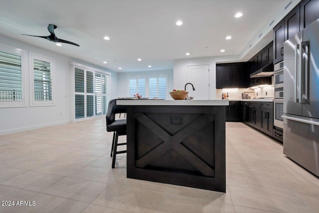 kitchen featuring ceiling fan, light tile patterned floors, a kitchen island with sink, stainless steel appliances, and a breakfast bar area