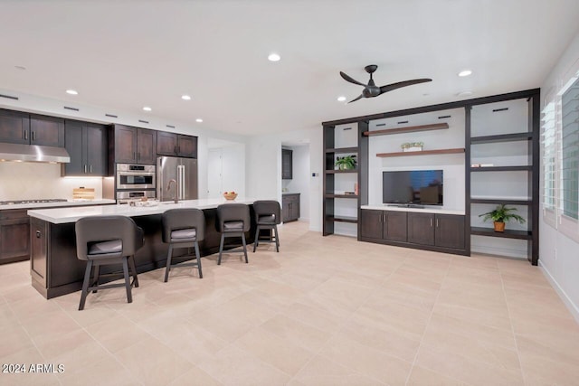 kitchen featuring a kitchen island with sink, stainless steel refrigerator, a kitchen breakfast bar, dark brown cabinetry, and ceiling fan