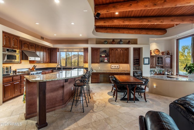 kitchen with a kitchen island with sink, under cabinet range hood, stainless steel oven, open shelves, and beamed ceiling