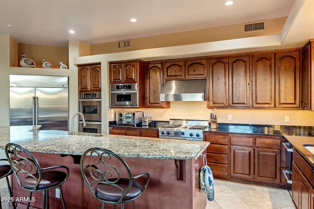 kitchen with light stone counters, stainless steel appliances, visible vents, an island with sink, and under cabinet range hood