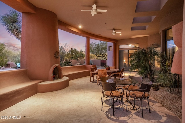 view of patio with a ceiling fan, outdoor dining space, and a lit fireplace