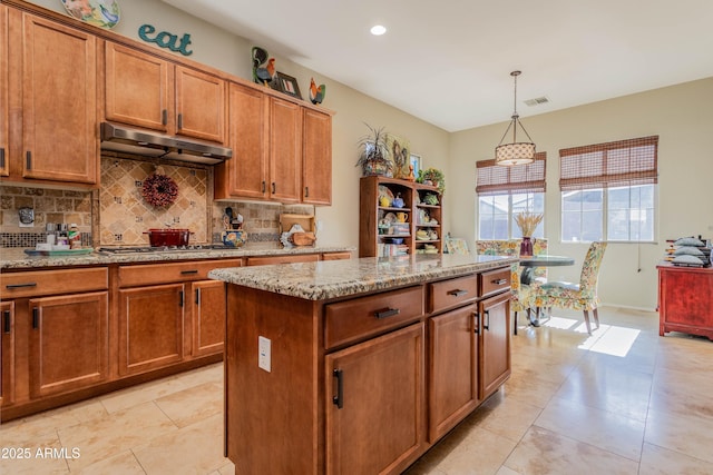 kitchen featuring exhaust hood, decorative light fixtures, a kitchen island, light stone counters, and stainless steel gas cooktop